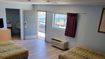 Habitación de hotel con dos camas, una ventana que da a un edificio, una puerta a un baño y un techo con una cámara de seguridad.