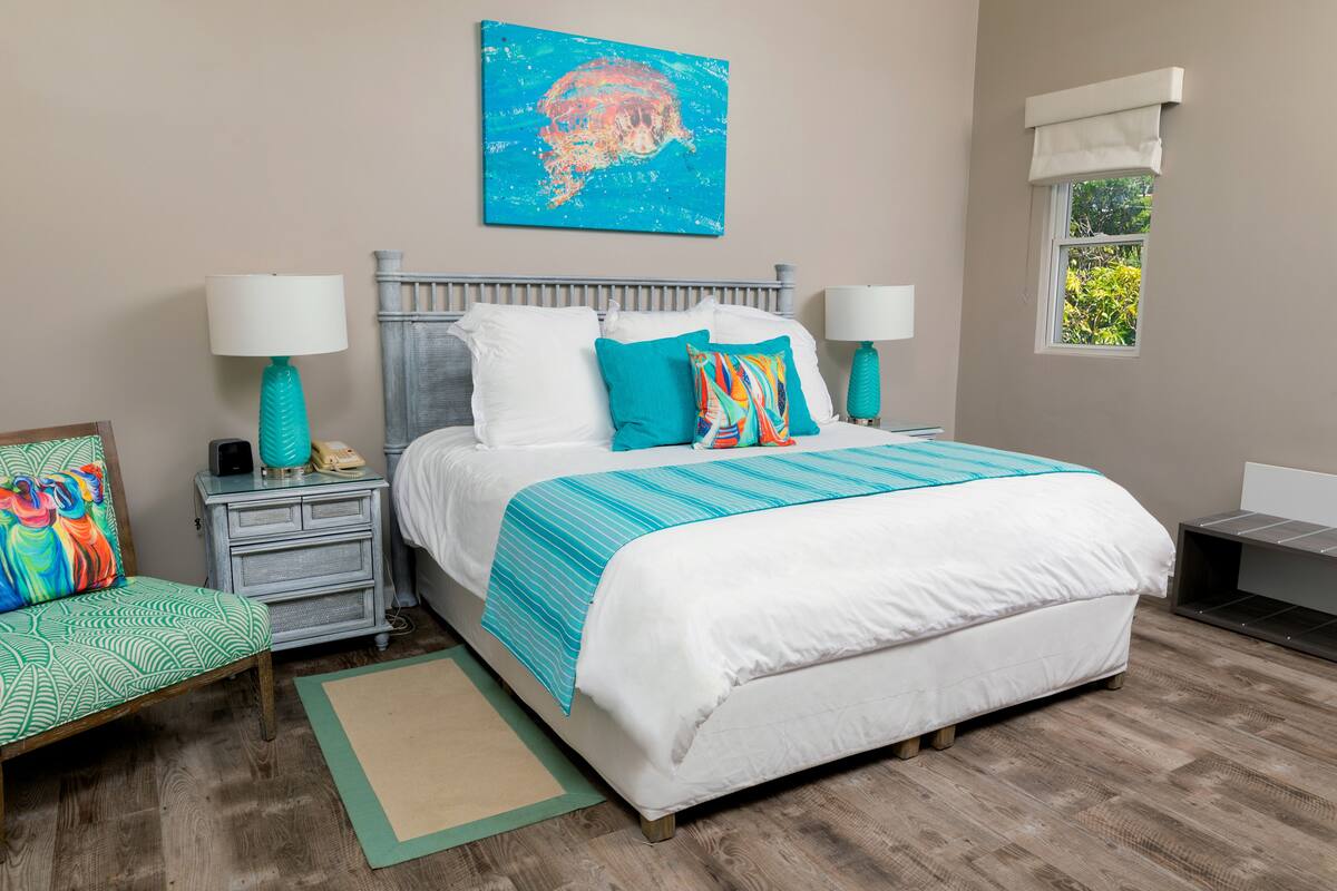 Une chambre avec un grand lit, deux tables de chevet, une commode, une fenêtre donnant sur un beau paysage et un tableau abstrait aux couleurs vives accroché au mur.