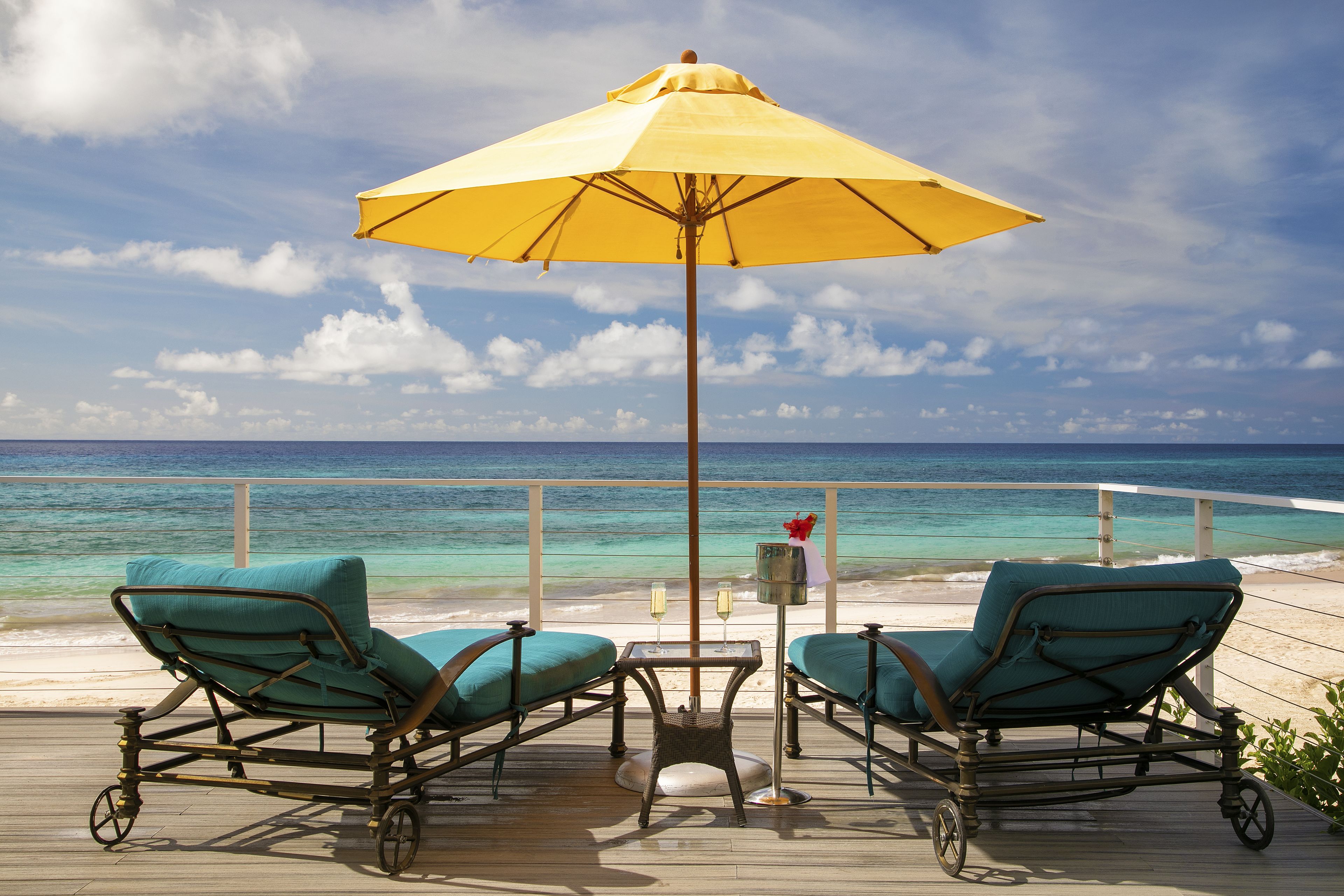 A deck with two lounge chairs and a table, a yellow umbrella, and a view of the ocean.