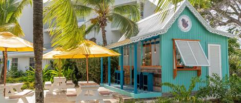 A turquoise building with a white roof and a balcony. There are outdoor seating areas with yellow umbrellas and white benches.