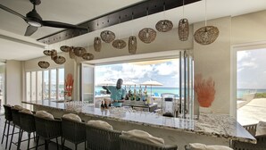 Un espace bar avec des chaises en osier, un comptoir équipé d’ustensiles de bar, et une vue sur l’océan grâce à de grandes fenêtres.