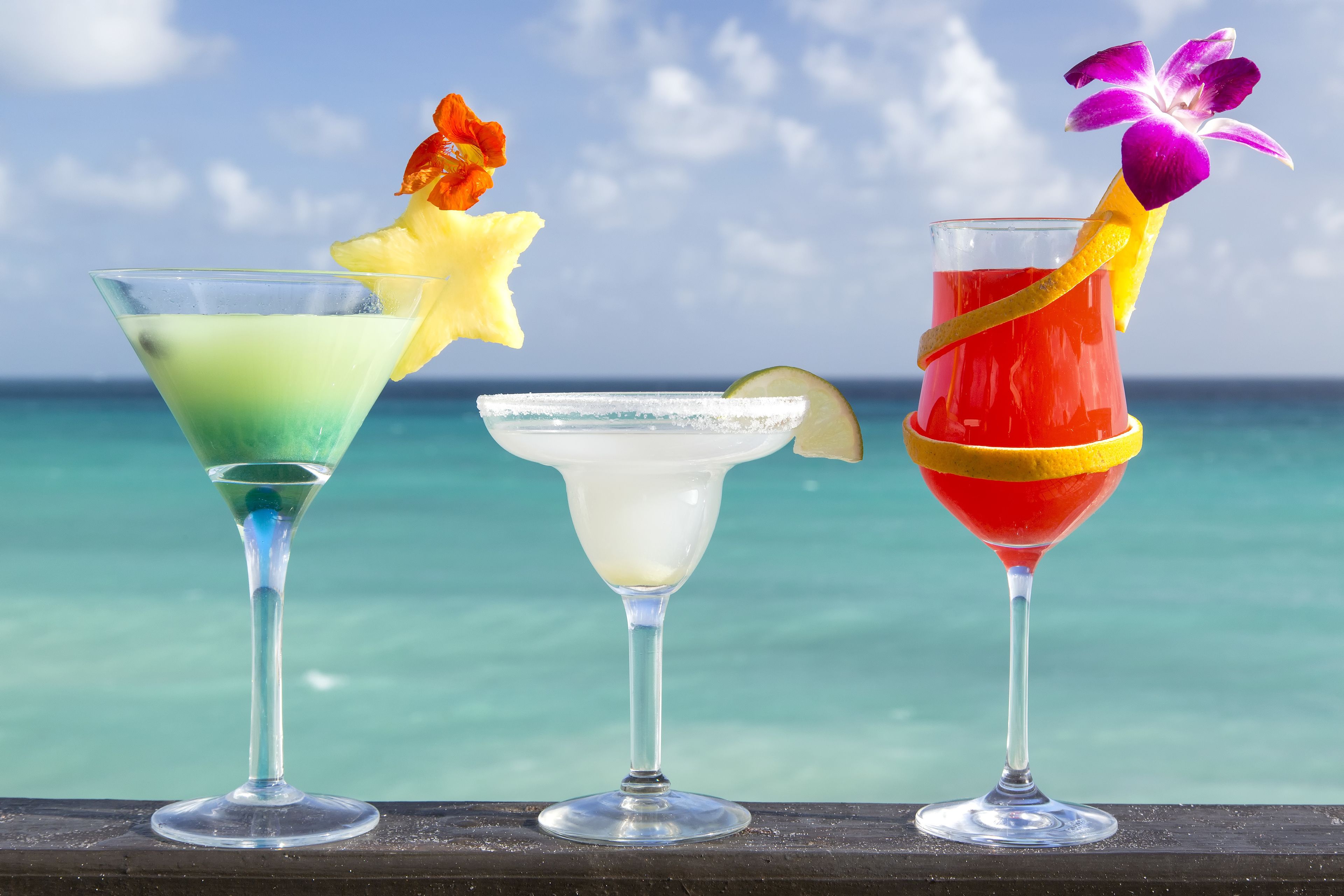 Three cocktails with tropical garnishes, set against a clear blue sky and ocean view.