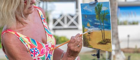 A person painting a landscape with palm trees and a sailboat.