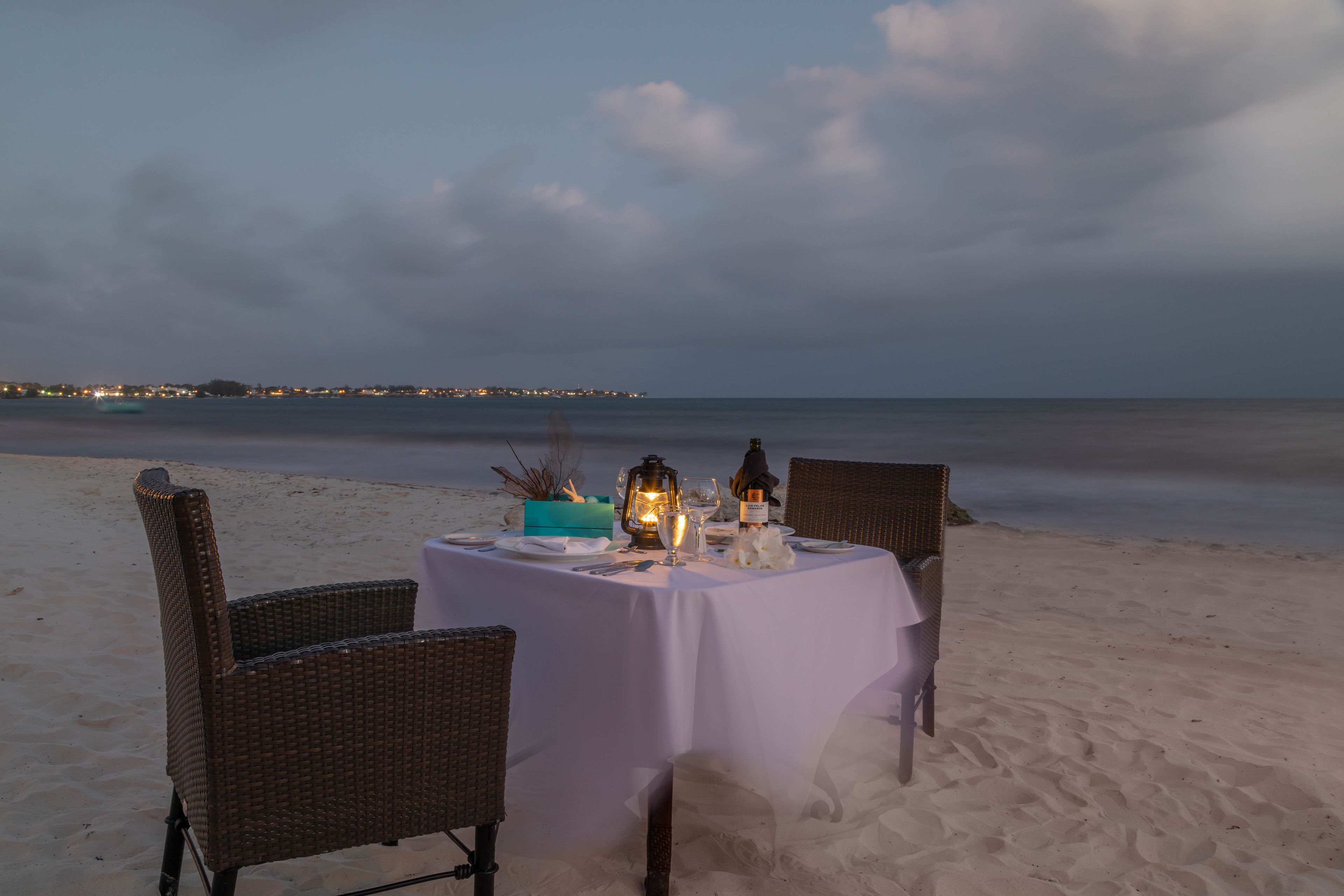 A table set for two on a beach with wicker chairs, a white tablecloth, and a view of the ocean.