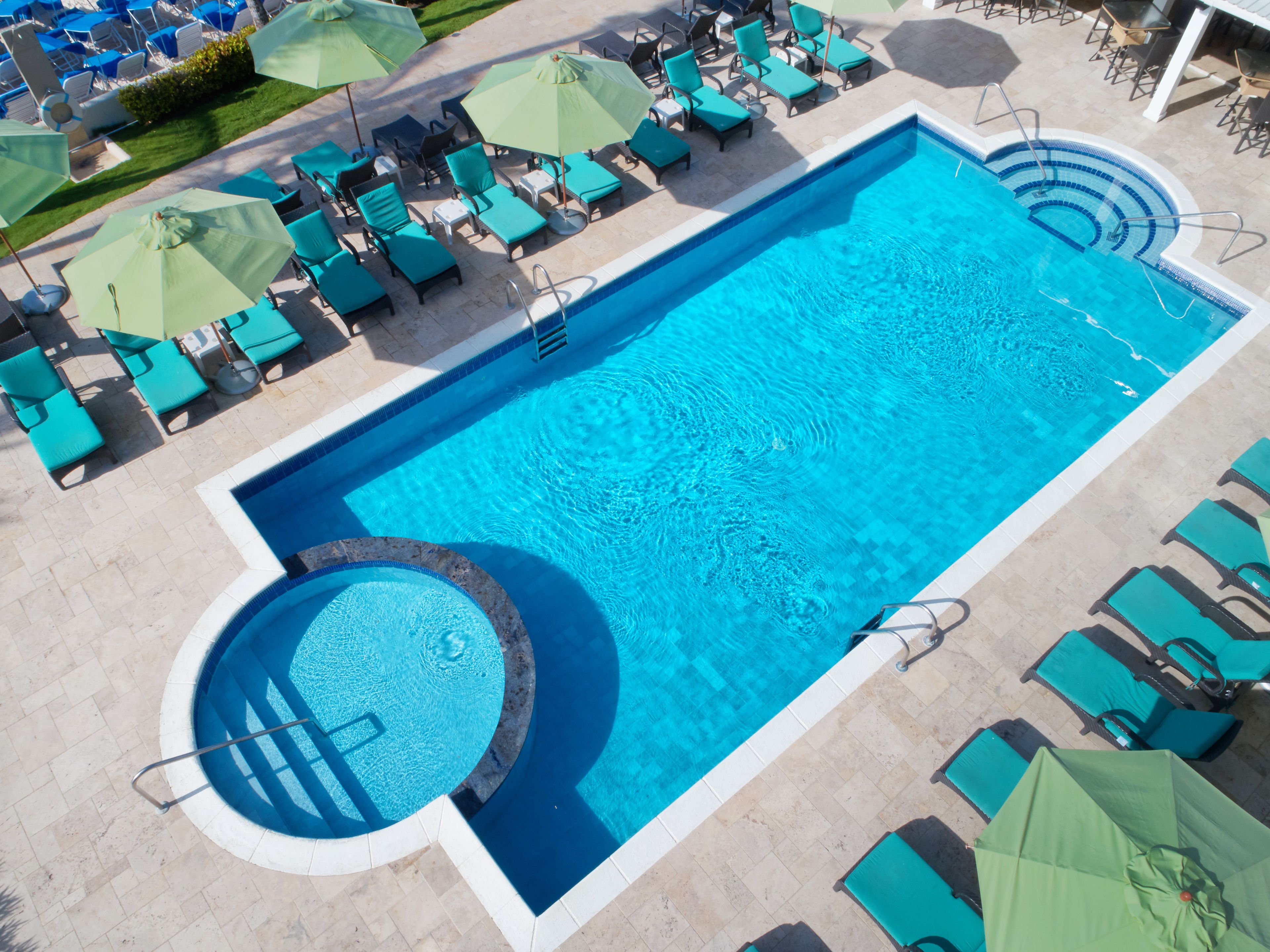 Una piscina rectangular grande con un jacuzzi circular, rodeada de numerosas tumbonas y sombrillas.