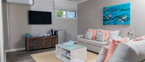 A modern living room with a sofa, coffee table, and TV. There is a painting on the wall and a window with blinds.
