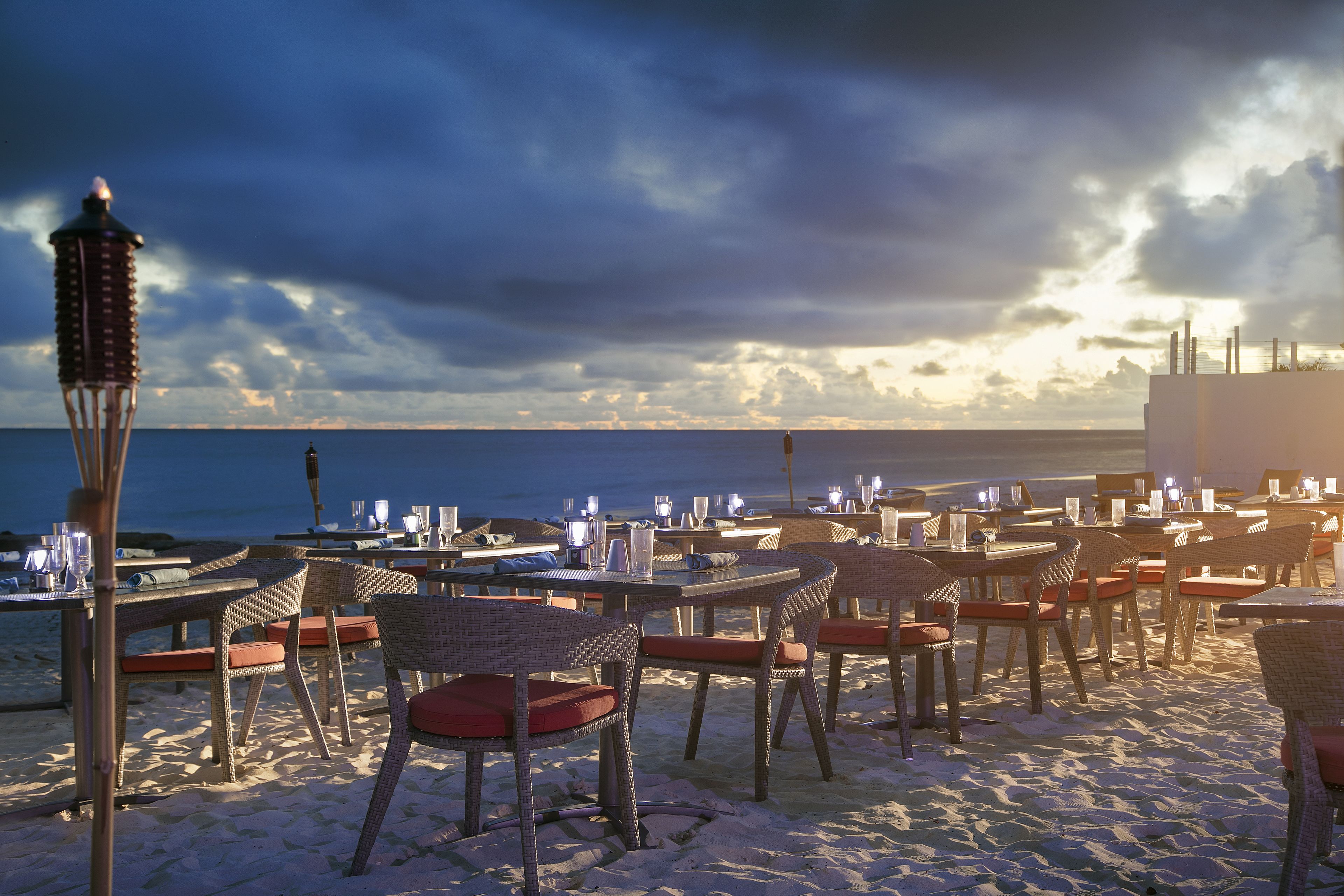 A beachside restaurant with wicker tables and chairs, a tiki lamp, and a view of the ocean.