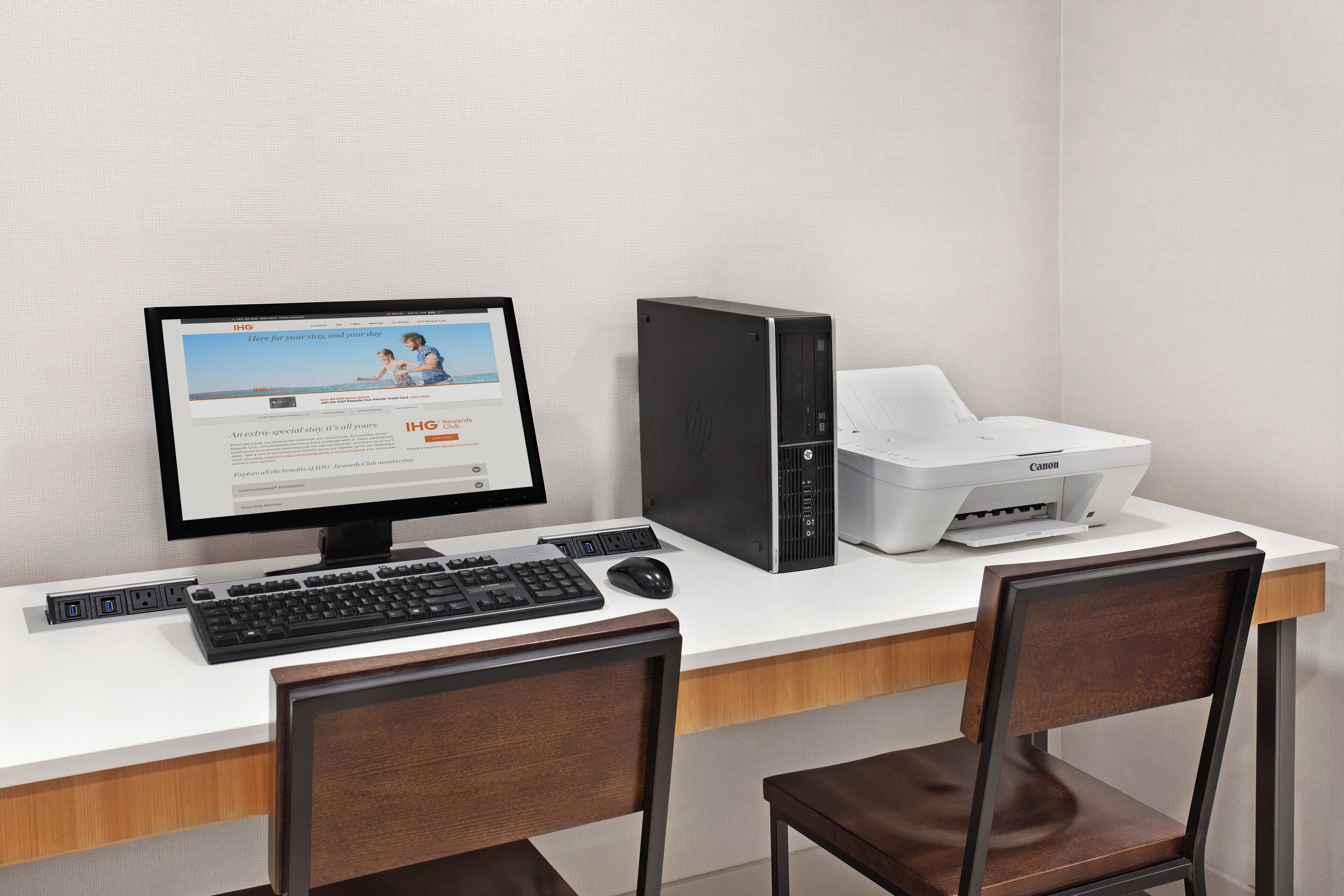 A hotel room with a desktop computer, a printer, and two chairs.