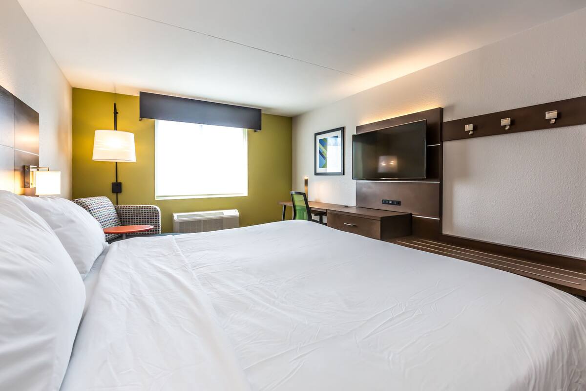 A hotel room with a large bed, a desk with a chair, a flat-screen TV, and a window with blinds.
