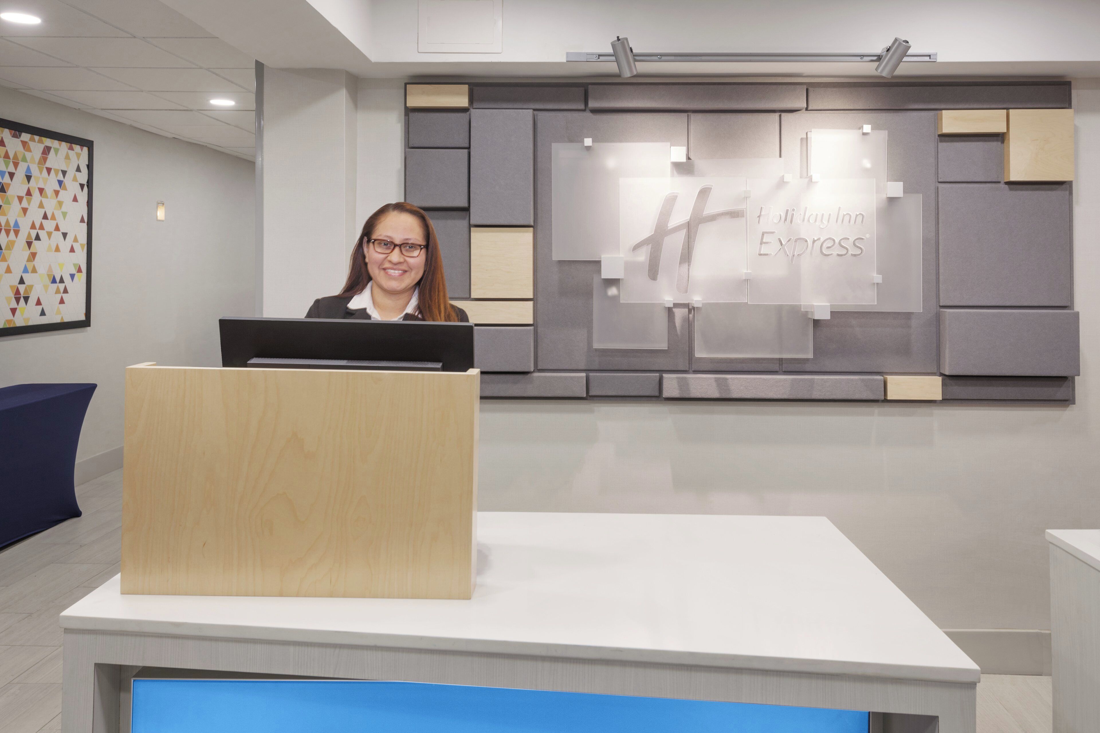 A reception desk with a computer, a wall with a #HolidayInn Express logo, and a colorful geometric wall art piece.