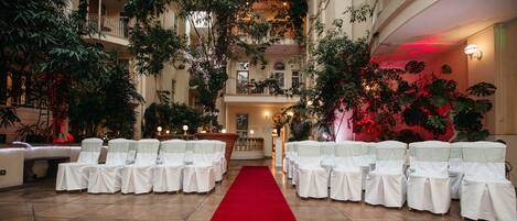 An indoor venue with a red carpet aisle, white chairs with sashes, and lush greenery.