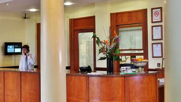 A hotel reception area with a large white column, a wooden reception desk, a chandelier, and a television screen.