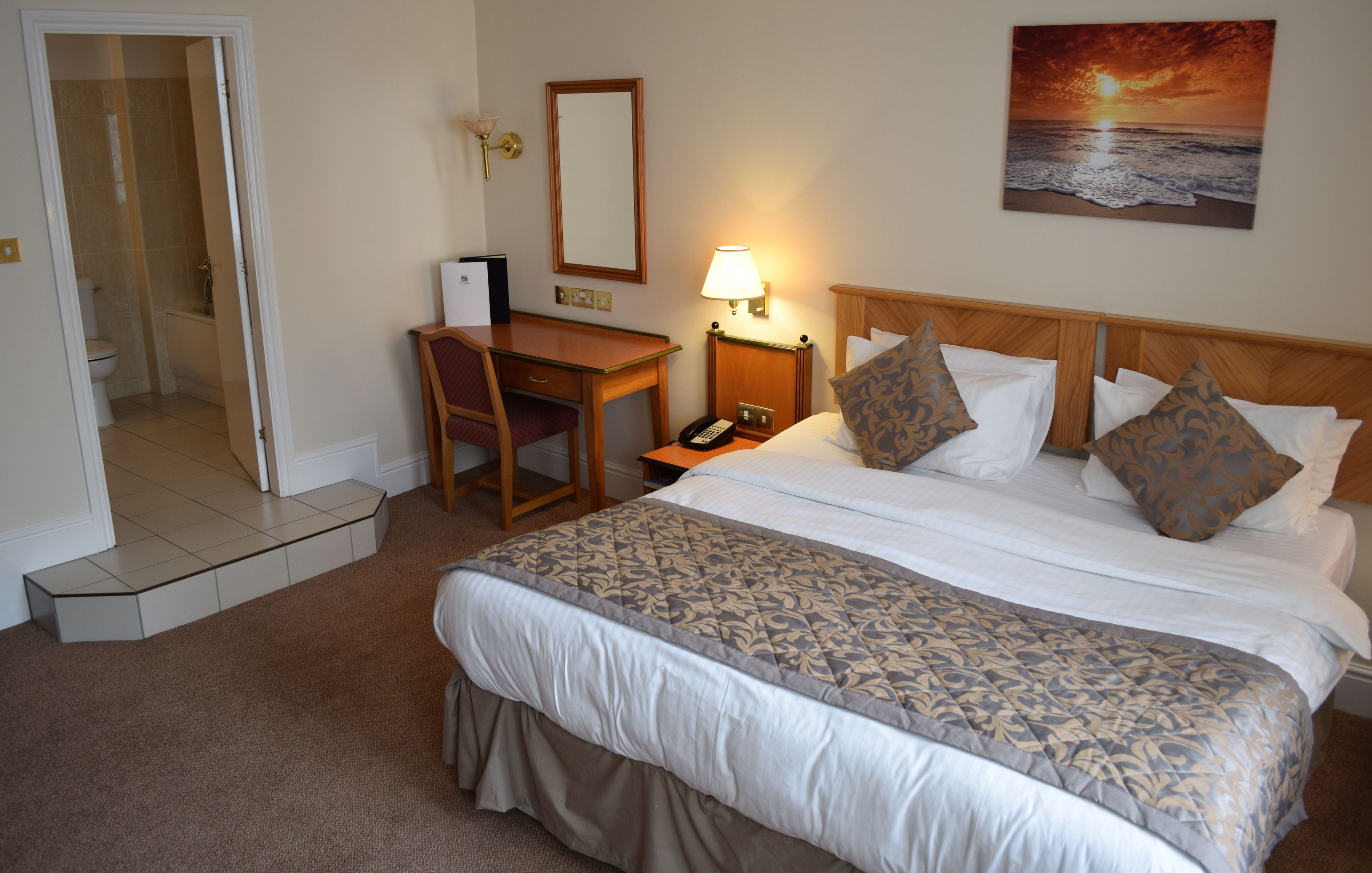 A hotel room with a bed, desk, chair, and a painting of a sunset over the ocean.