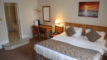 A hotel room with a bed, desk, chair, and a painting of a sunset over the ocean.