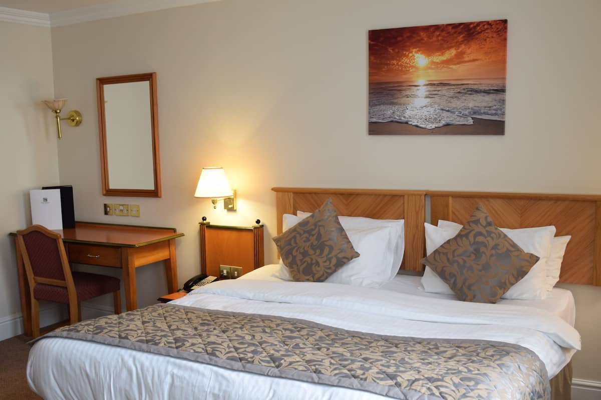 A hotel room with a large bed, a desk, a chair, a lamp, and a painting of a sunset over the ocean.