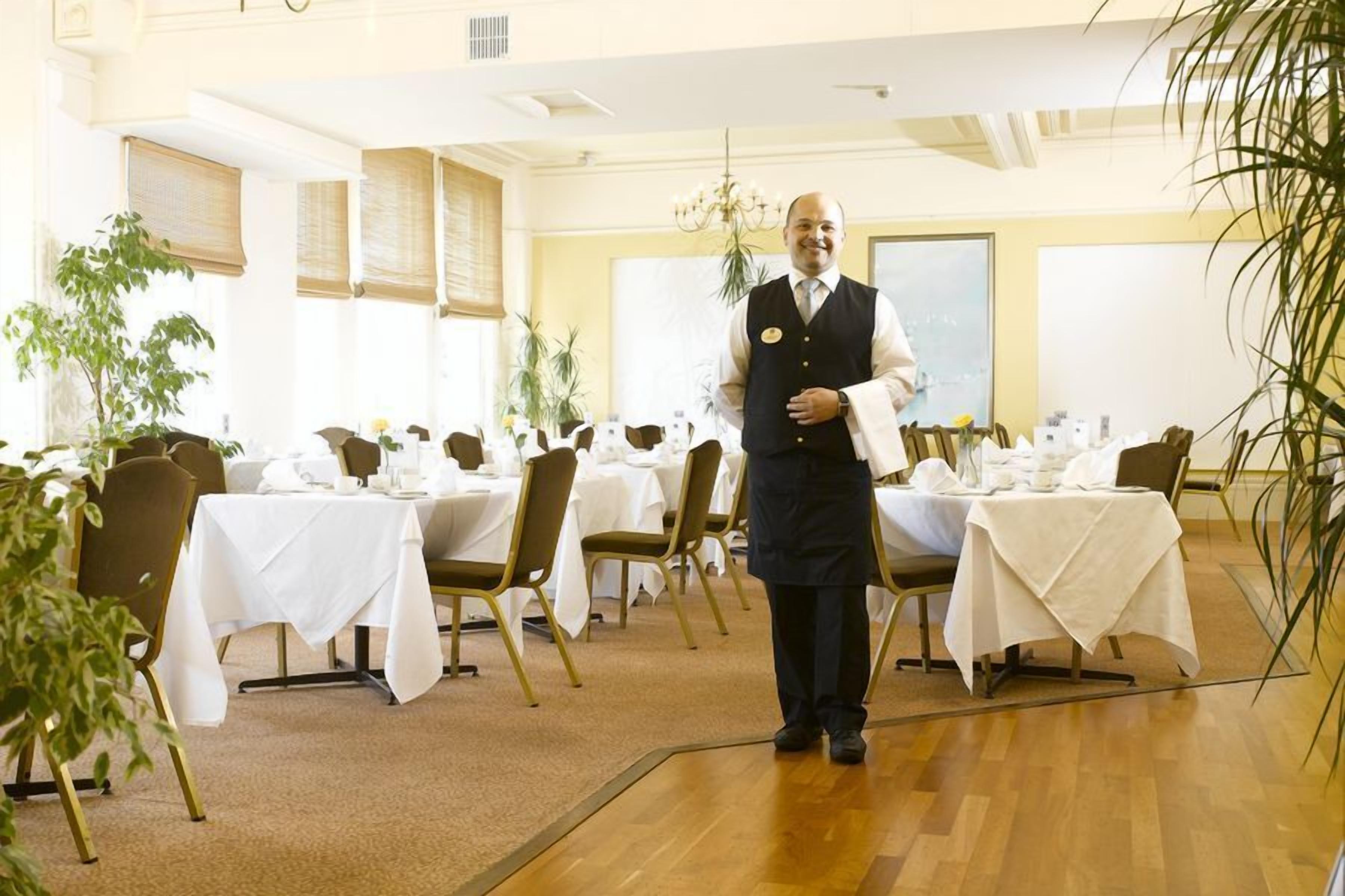 Un comedor de hotel con varias mesas preparadas para el servicio, un camarero con uniforme y plantas en macetas.