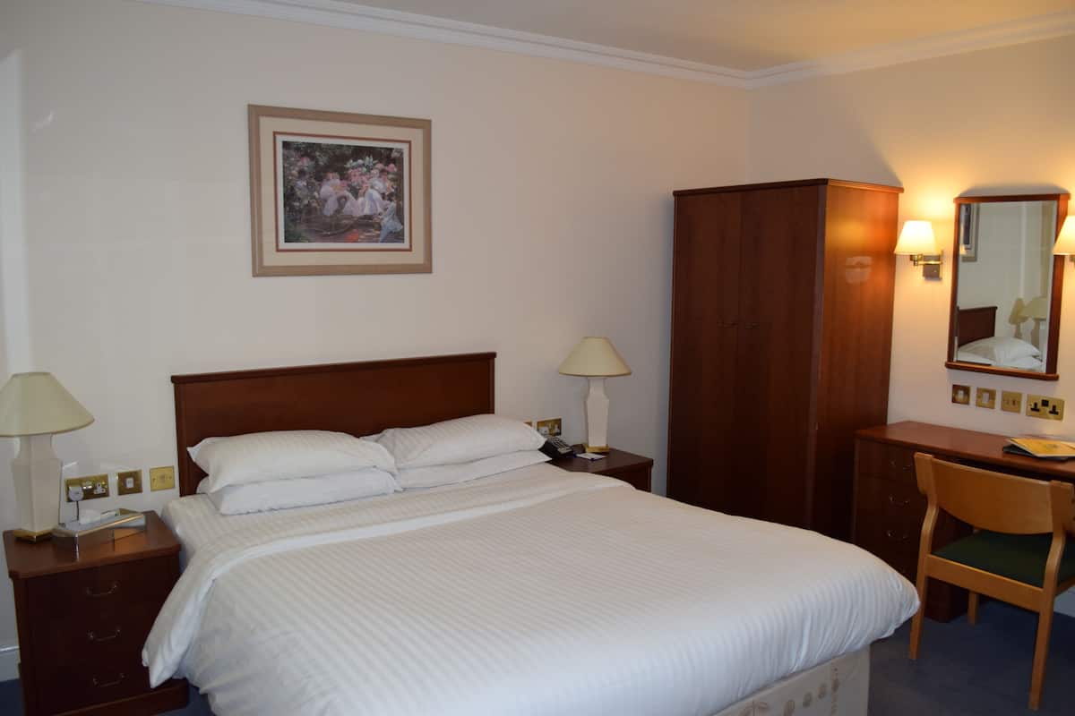 A neatly made bed with white linens, a wooden nightstand with a lamp, a wooden wardrobe, a desk with a chair, and a framed picture on the wall.