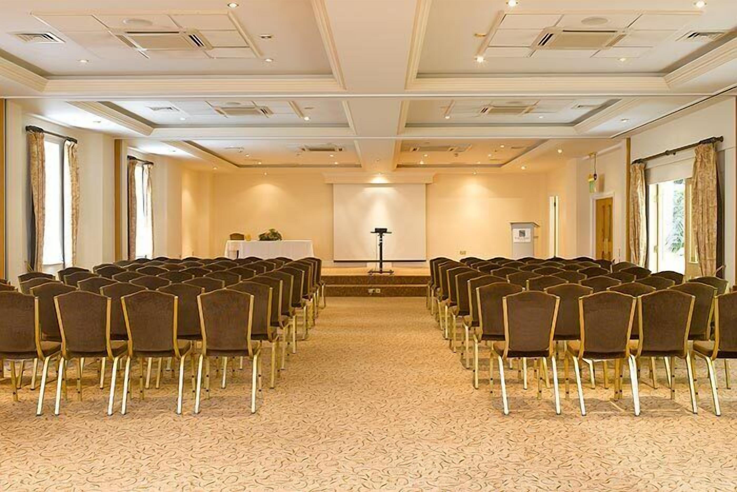 Een vergaderzaal met rijen stoelen gericht op een spreekgestoelte en een projectiescherm.