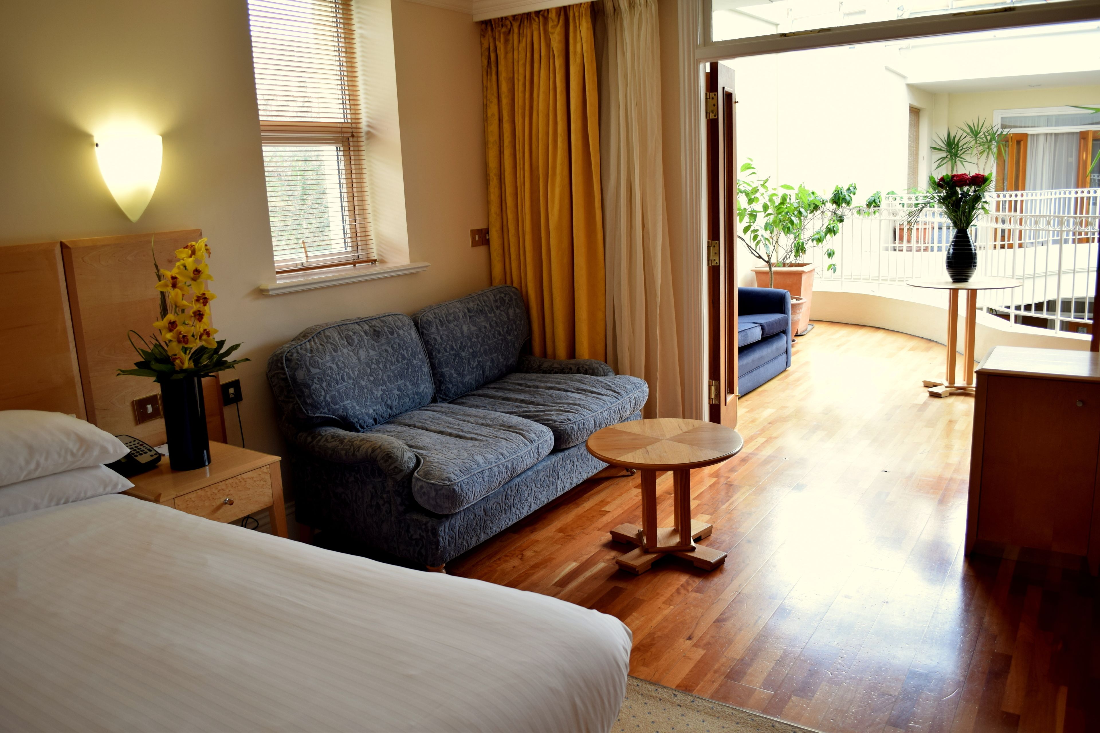 A hotel room with a bed, a sofa, a small table, and a balcony with plants.