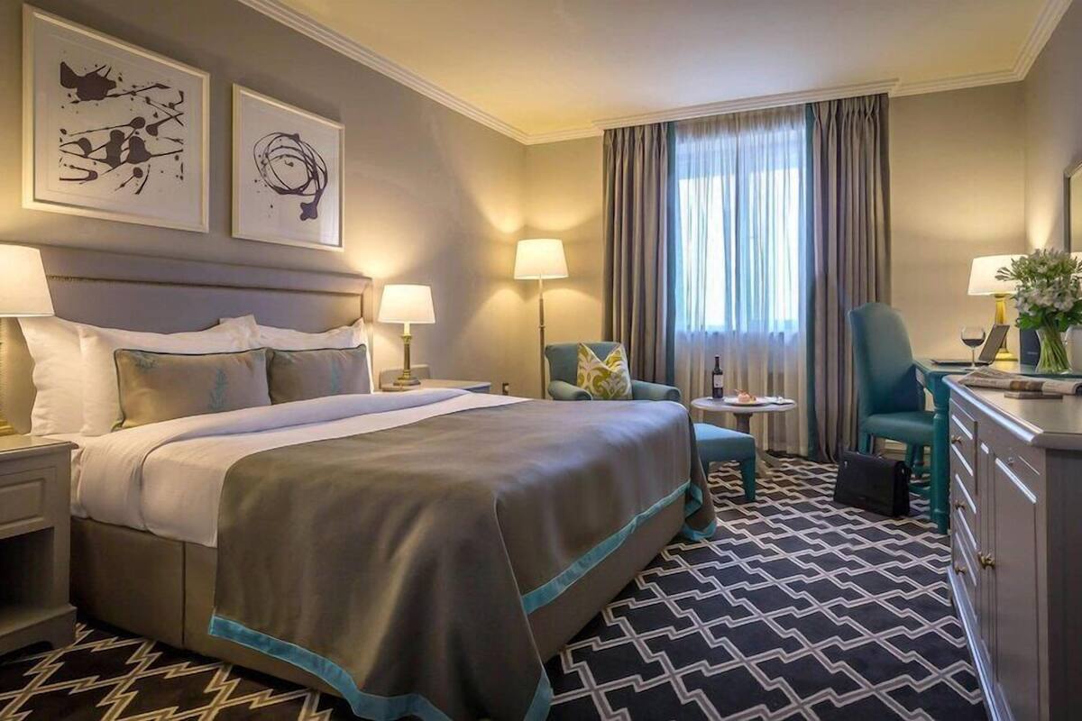 Une chambre d’hôtel avec un grand lit, deux tables de chevet avec des lampes, un bureau avec une chaise, une petite table ronde avec une chaise, un miroir et des œuvres d’art accrochées au mur.