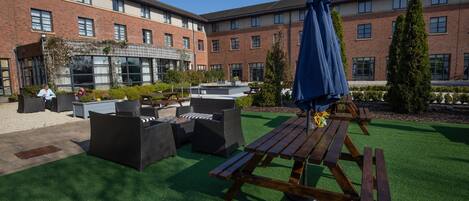 An outdoor patio with artificial grass, wooden picnic tables, and wicker furniture.