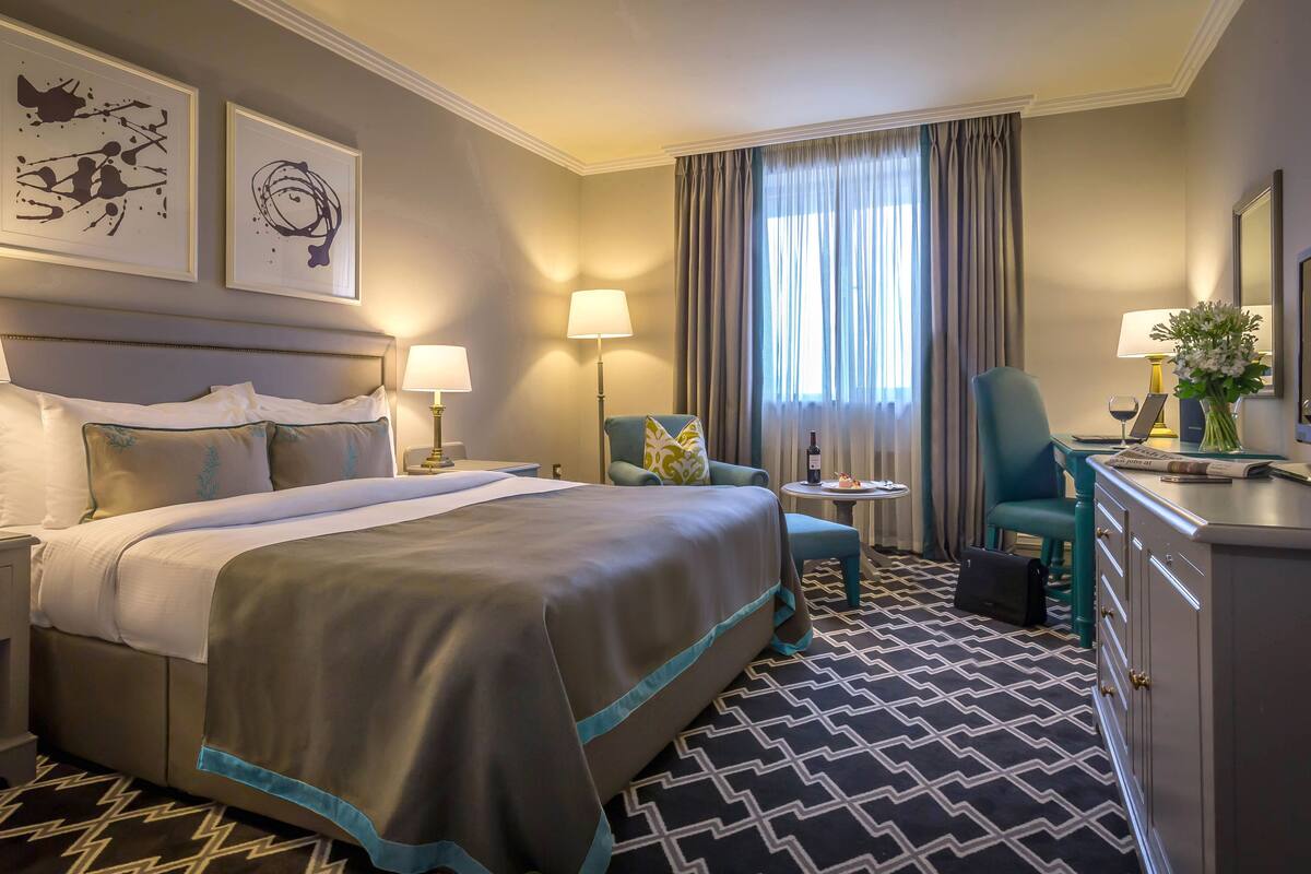 Une chambre d’hôtel avec un grand lit, deux tables de chevet, un bureau avec une chaise, une petite table ronde avec une lampe et un miroir.