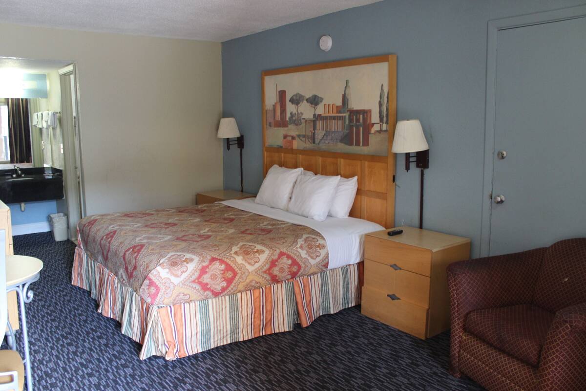 Ein Hotelzimmer mit einem Bett, Nachttischen, einem Schreibtisch, einem Sessel und einem Bild an der Wand sowie einer Tür.