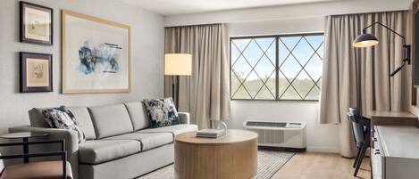 A modern living room with a grey sofa, a round coffee table, and a view of the outdoors through large windows.