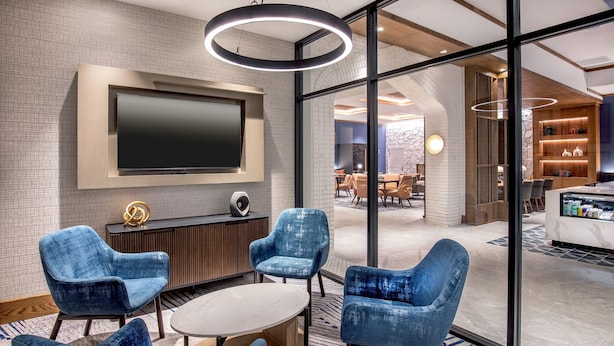 A modern lounge area with blue upholstered chairs, a round table, a flat-screen TV mounted on the wall, and a glass partition leading to a dining area.