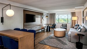 A modern hotel room with a wooden dining table, blue chairs, a round coffee table, a sofa, and a TV mounted on the wall.