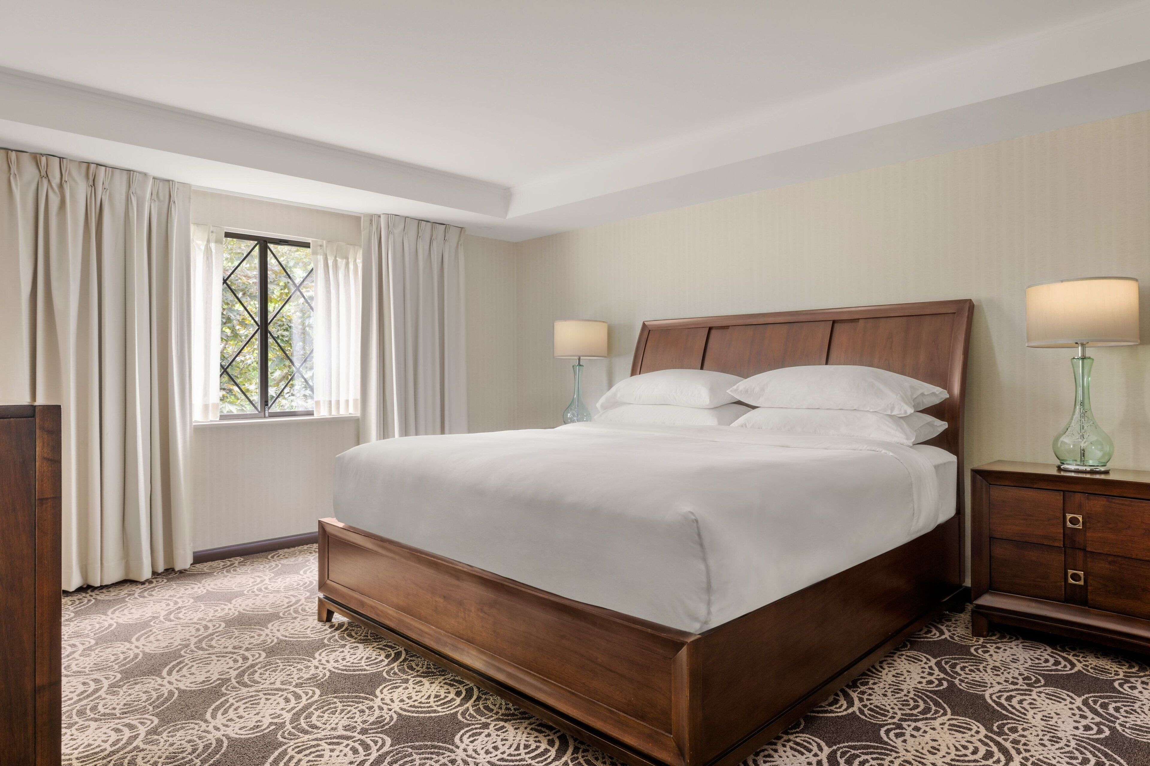 A hotel room with a large bed, two bedside tables, a window with curtains, and a patterned carpet.