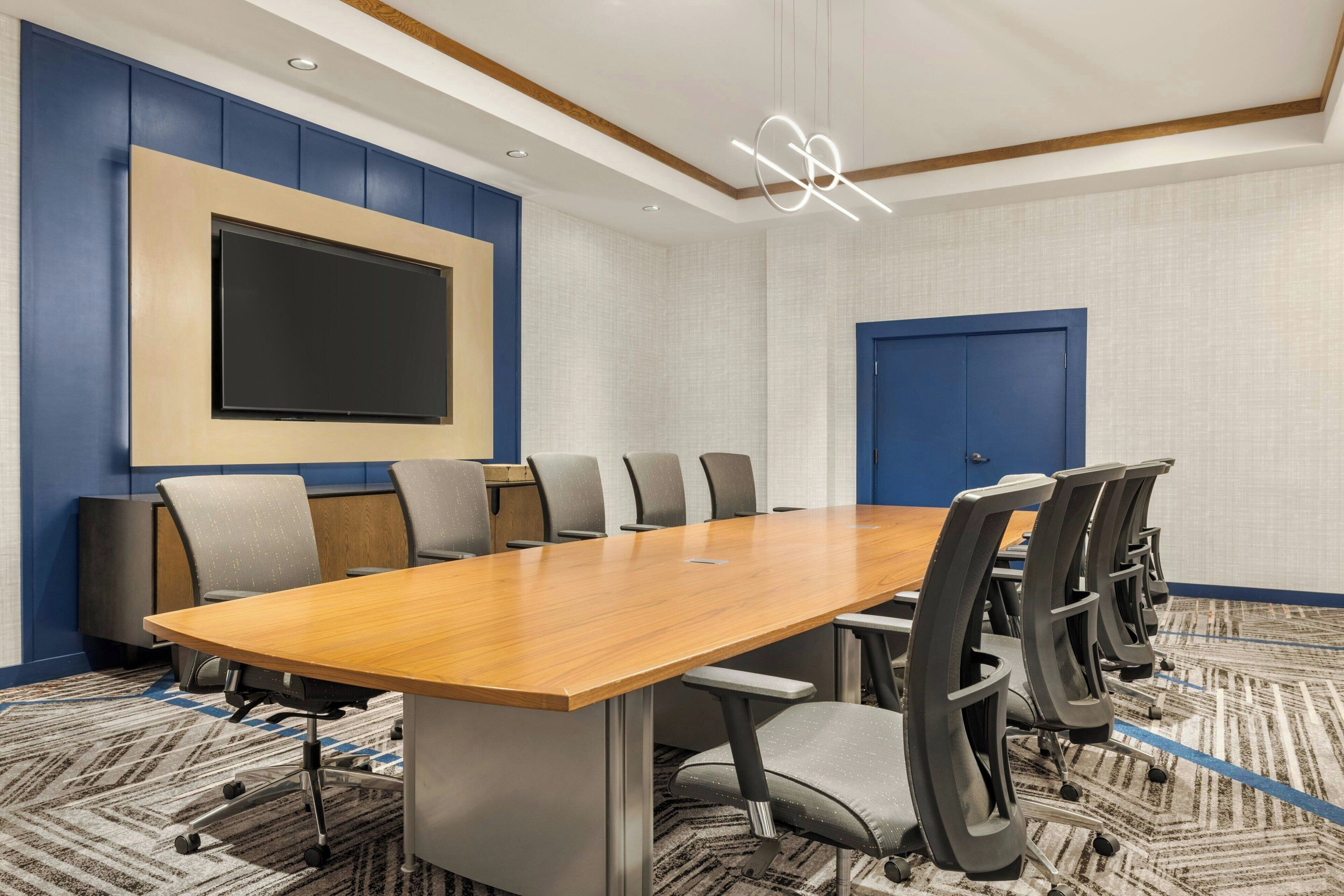 A conference room with a long wooden table, multiple chairs, a large flat-screen TV mounted on the wall, and a blue door.