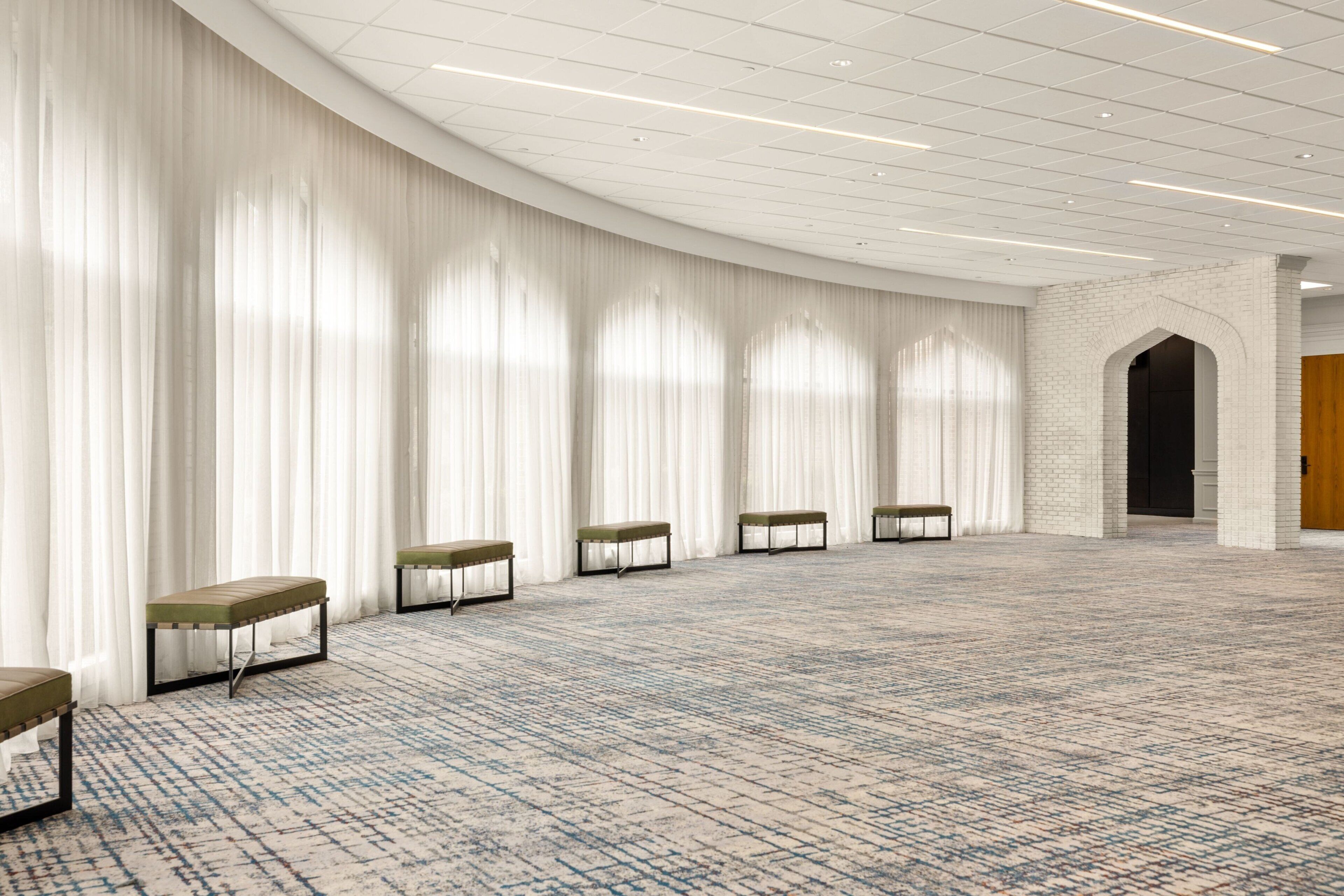 A spacious room with a patterned carpet, white curtains, and a series of benches along the wall.