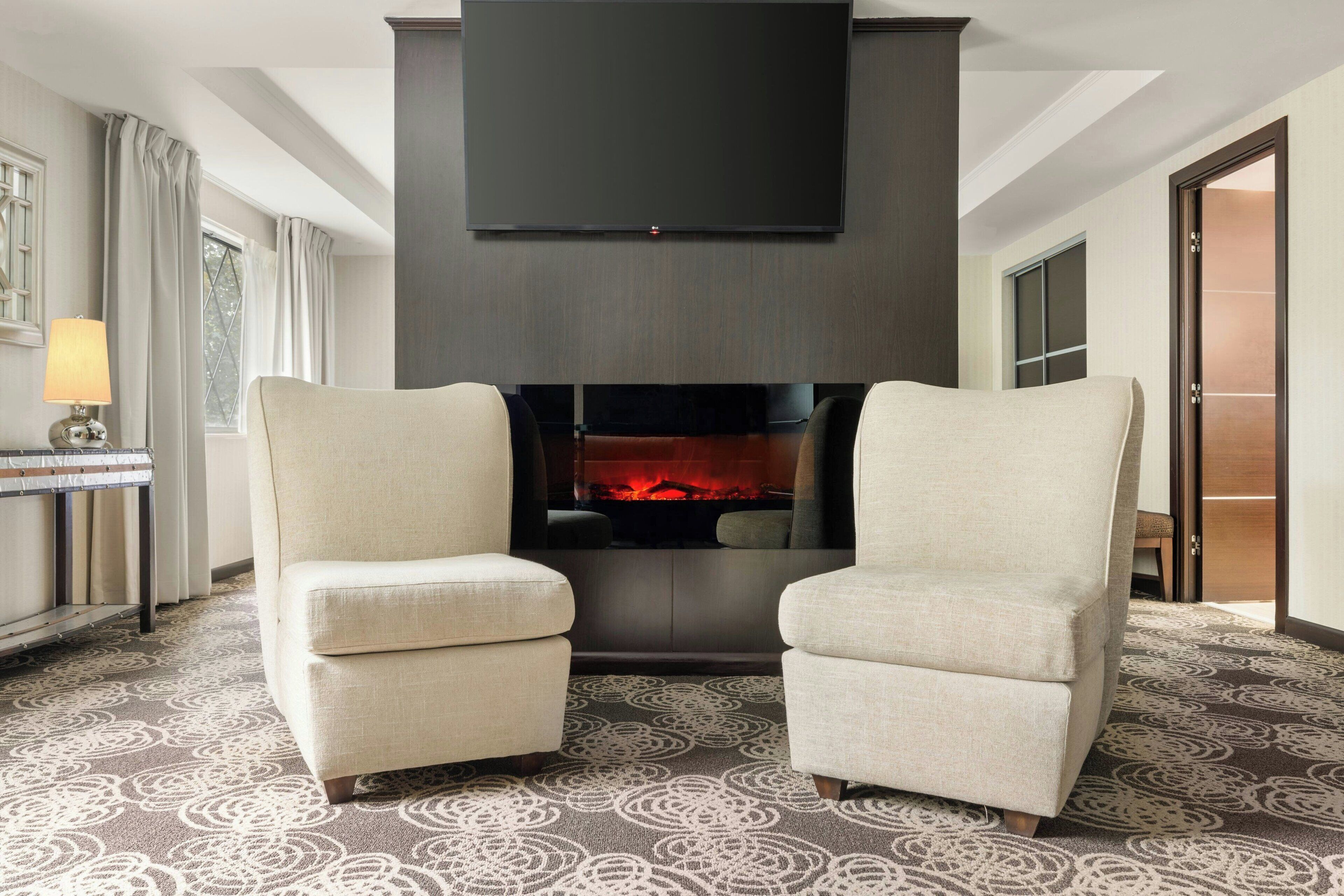 A hotel room with a fireplace, two armchairs, a TV mounted on the wall, and a patterned carpet.