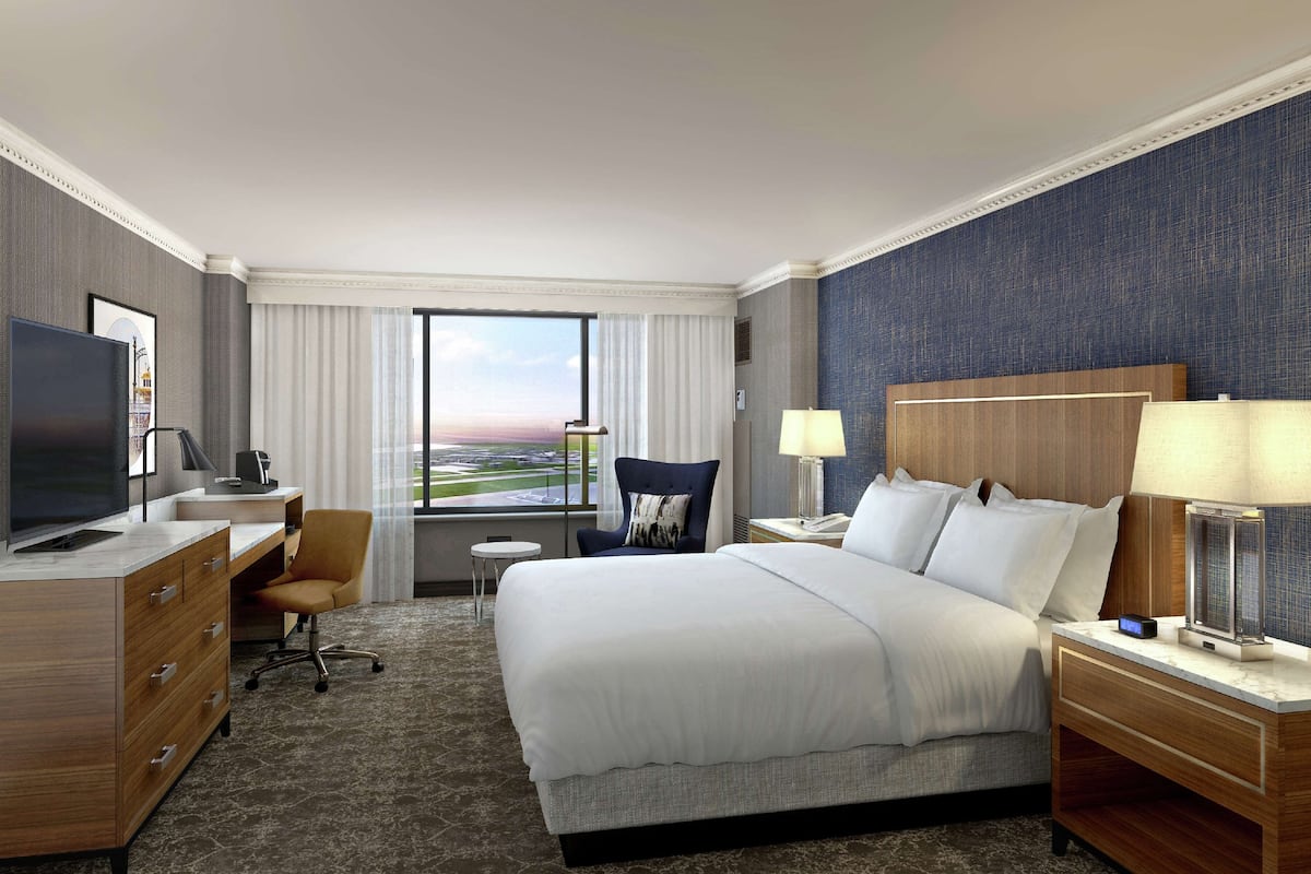Hotelkamer met een groot bed, een bureau met een televisie, een stoel, een tafeltje en uitzicht op het vliegveld.