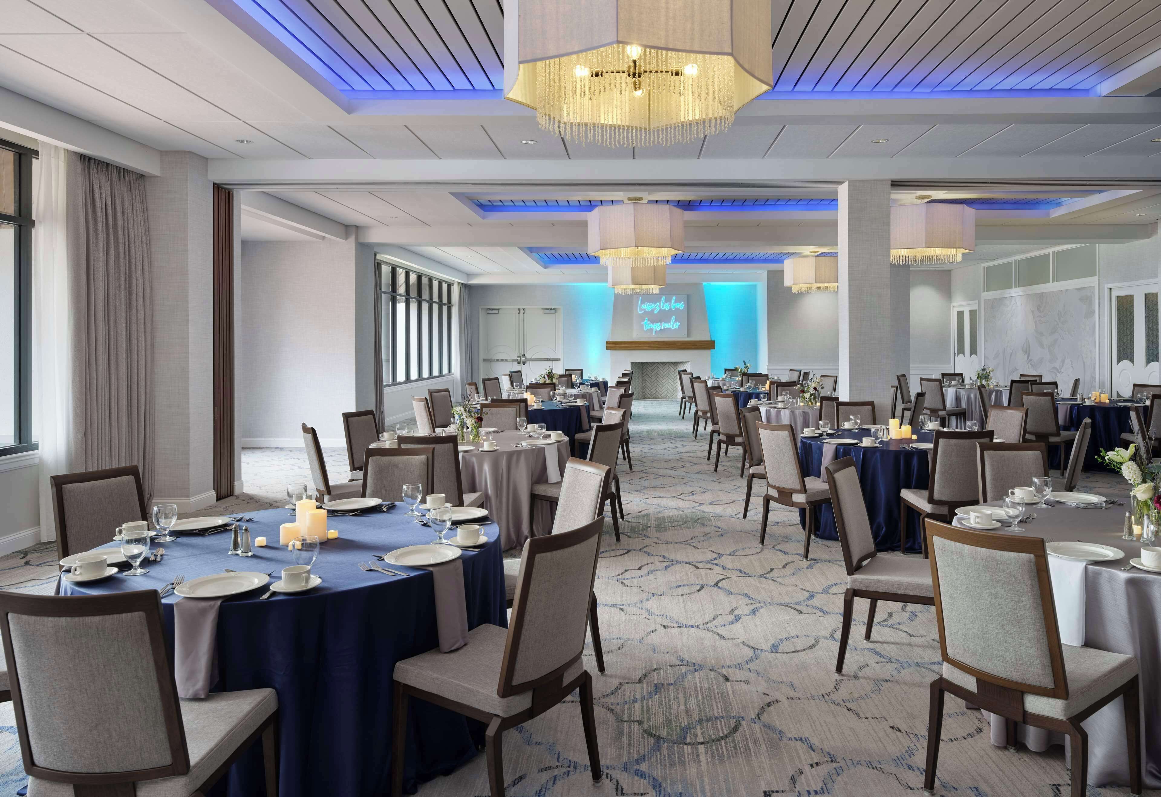 A spacious conference room with round tables set for dining, a large screen at the front, and a modern chandelier.