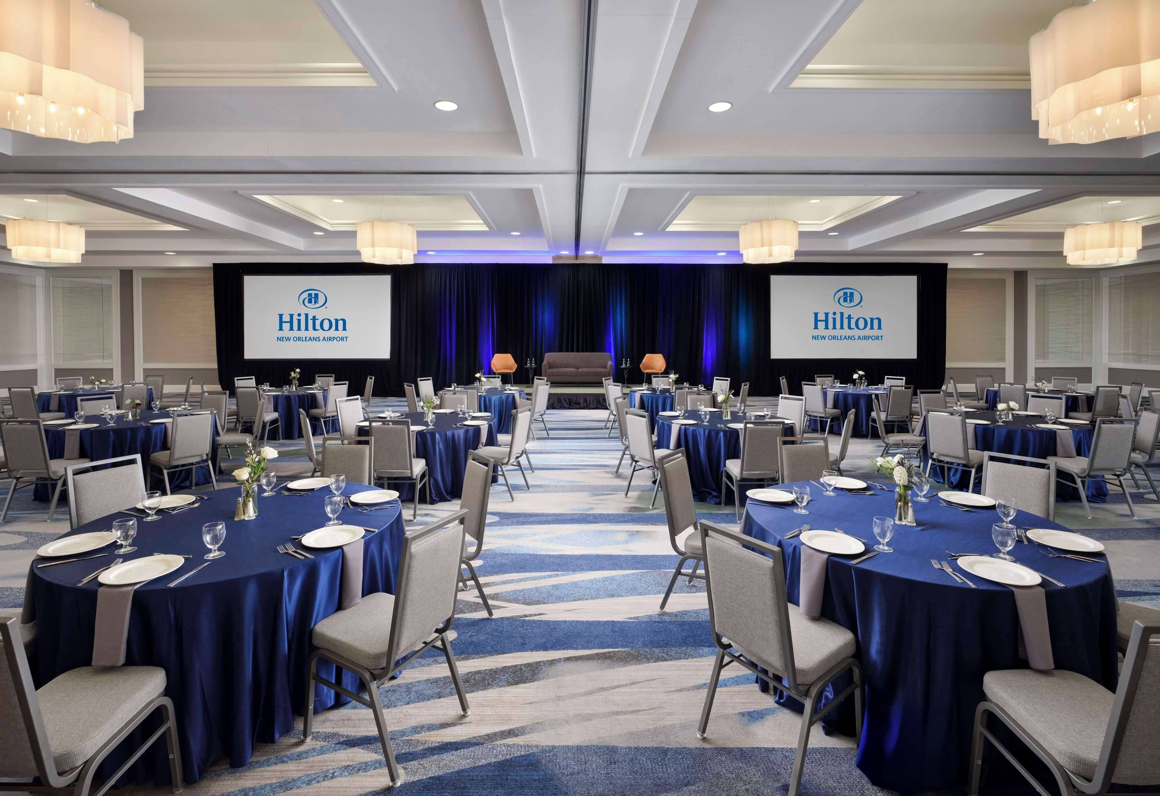 Une salle de conférence équipée de tables rondes, de chaises et d’une scène sur laquelle est installé un écran affichant le logo de Hilton.