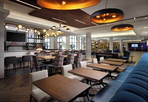 Een moderne hotellobby met een bar, zitgedeeltes en een groot scherm waarop het Hilton-logo staat.