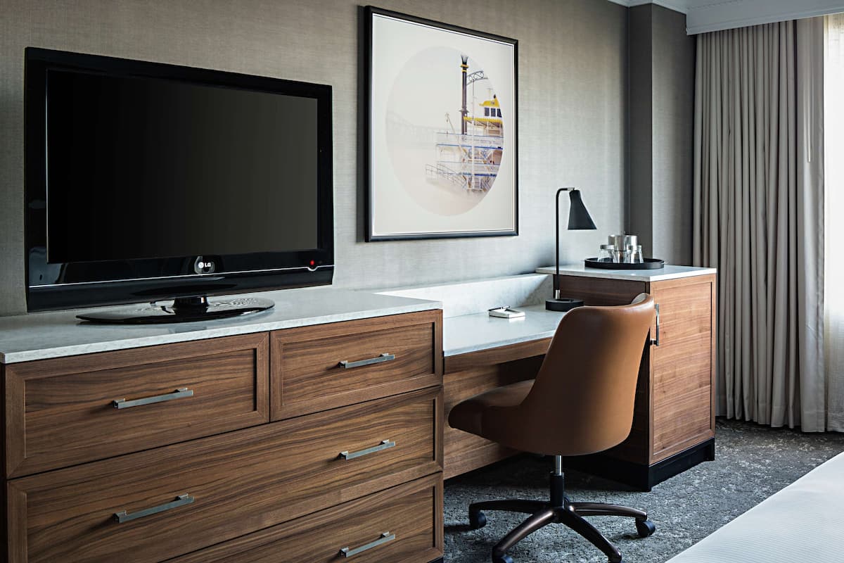 Een hotelkamer met een grote flatscreen televisie, een bureau met een computer, een stoel, een kast en een lijst met een afbeelding aan de muur.