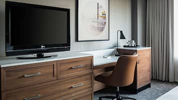A hotel room with a large flat-screen TV, a desk with a computer, a chair, a cabinet, and a framed picture on the wall.