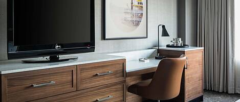 A hotel room with a large flat-screen TV, a desk with a computer, a chair, a cabinet, and a framed picture on the wall.