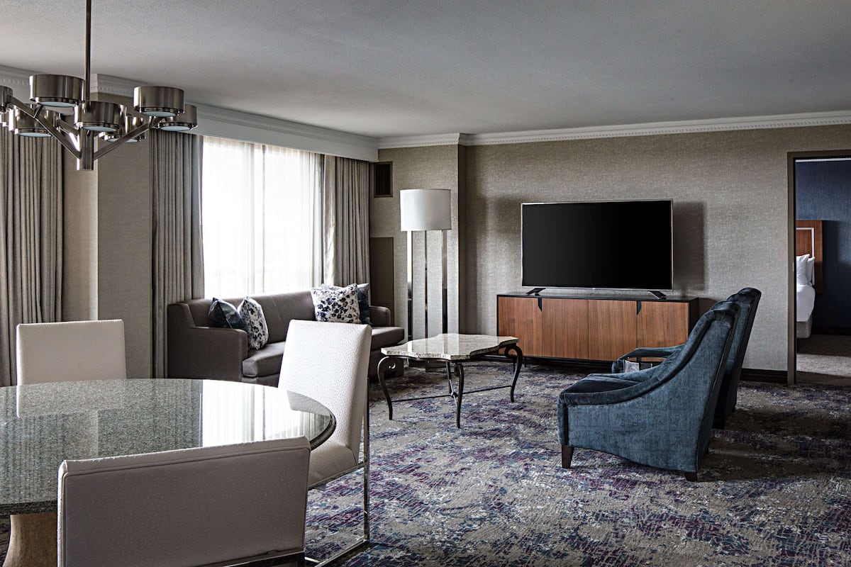 Een hotelkamer met een modern interieur, inclusief een bank, fauteuils, een eettafel, een flatscreen-tv en een groot raam met gordijnen.