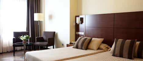 Habitación de hotel con una cama grande, dos sillones, una mesita con un florero y una lámpara.