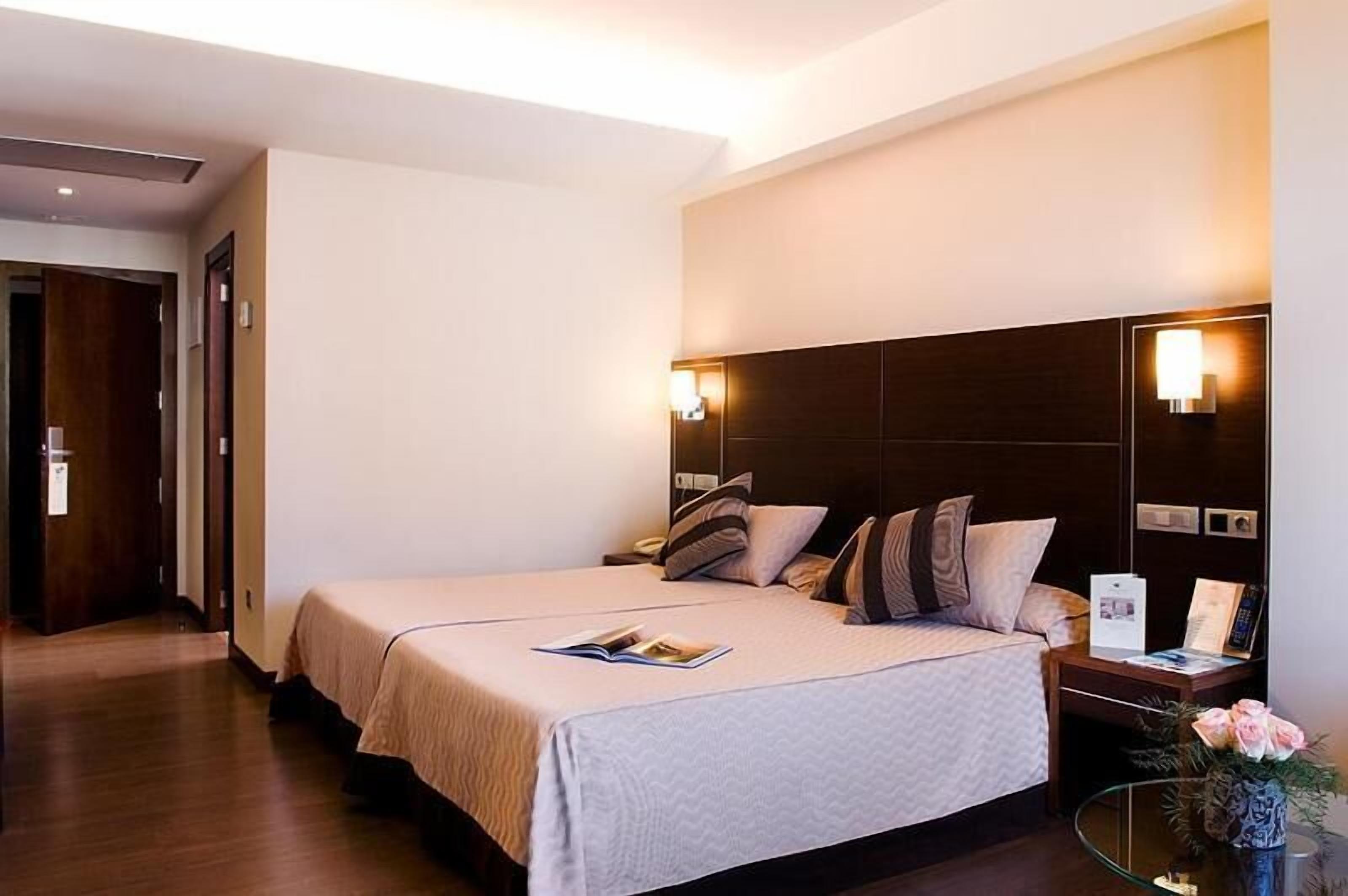 A hotel room with a large bed, bedside tables, a nightstand, a mirror, and a small table with a vase of flowers.