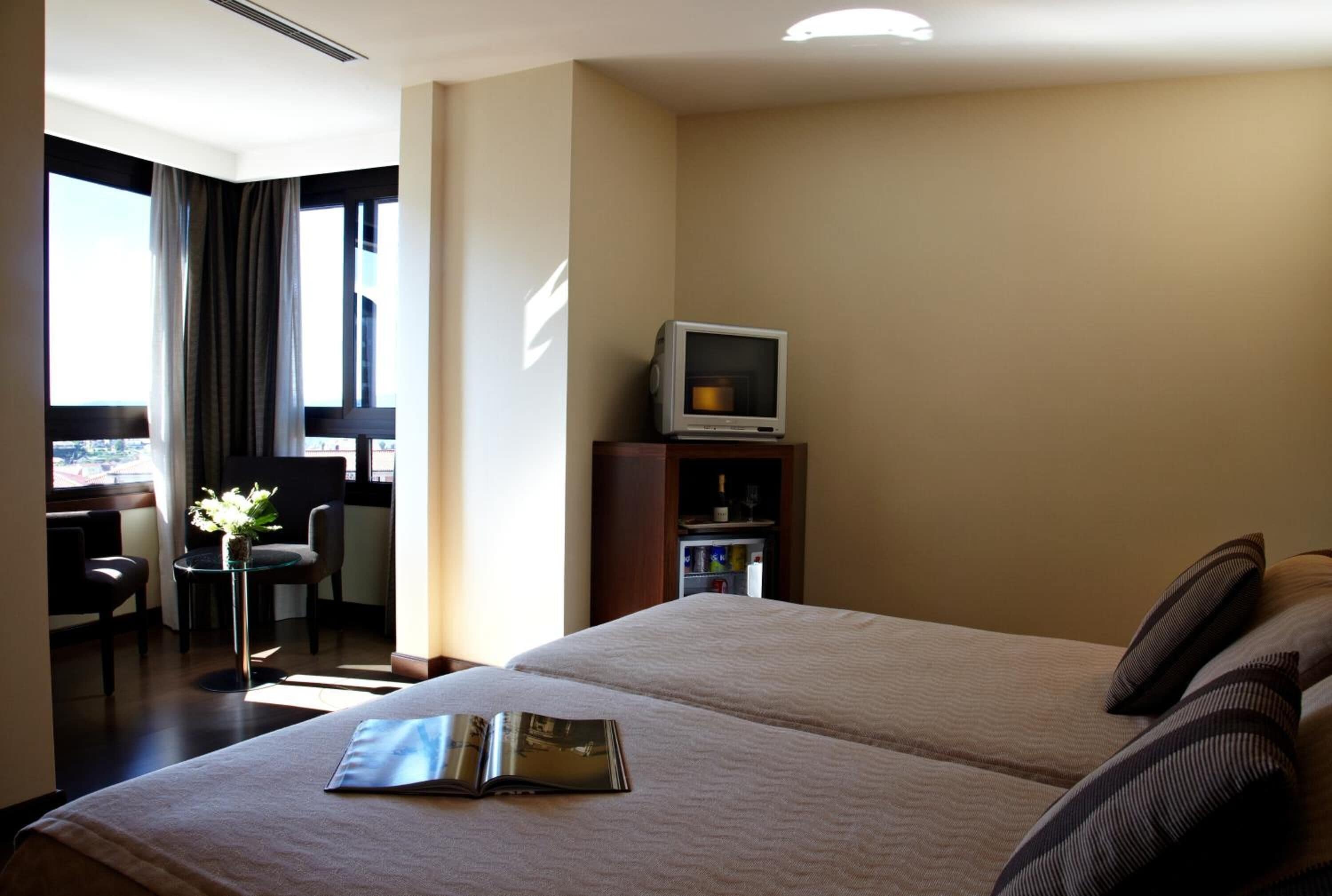 Habitación de hotel con dos camas, un televisor pequeño sobre un mueble, un escritorio con una silla y una ventana con cortinas.
