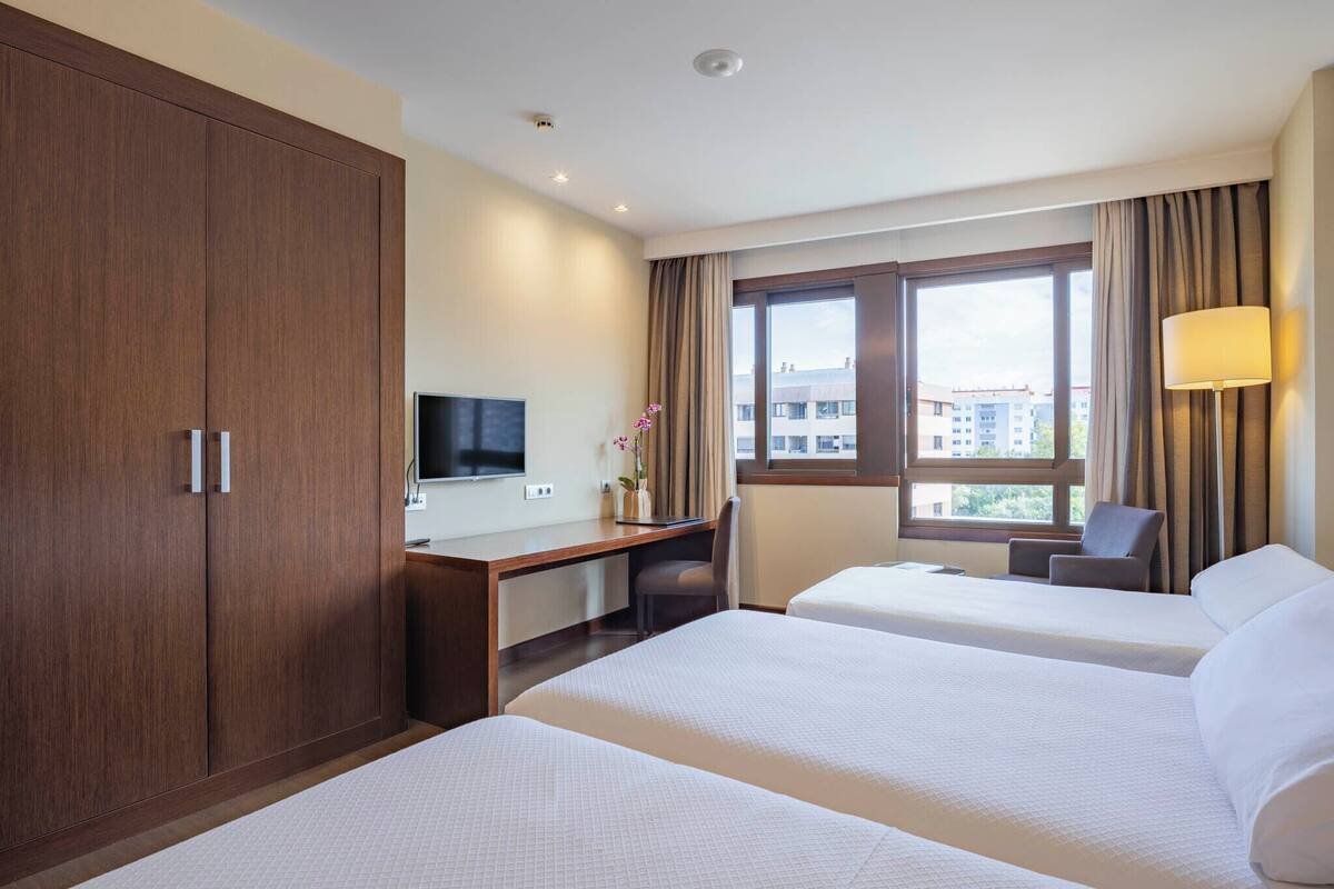 Een hotelkamer met twee bedden, een bureau, een televisie en een raam met uitzicht op gebouwen.