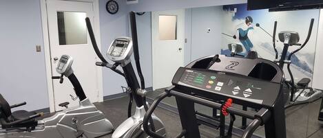 A gym with various exercise machines, including treadmills and elliptical trainers, with a large mirror and a television mounted on the wall.