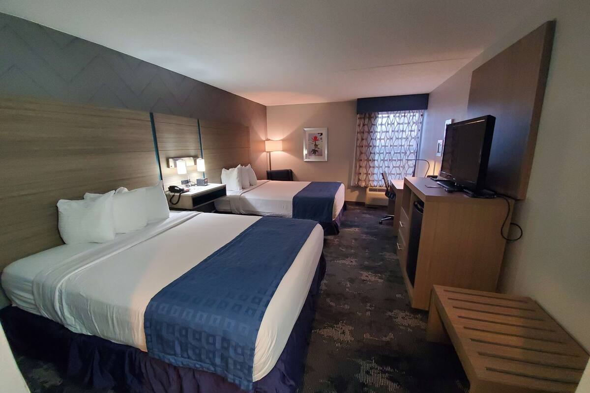 Quarto de hotel com duas camas, uma escrivaninha, uma televisão e uma janela com cortinas estampadas.