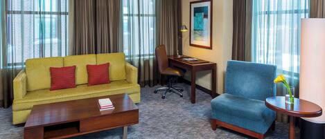 Une chambre d’hôtel avec un canapé jaune, un fauteuil bleu, une table basse en bois, un bureau avec une lampe et un tableau encadré au mur.