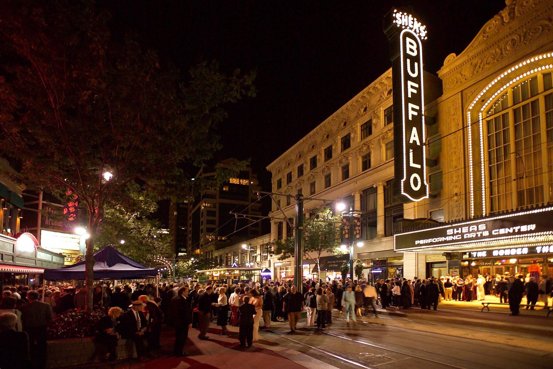 A busy street scene at night with a prominent theater marquee reading "BUFFALO" and "SHEAS PERFORMING ARTS."